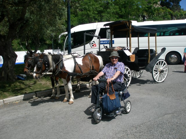A smiling man sat on his Di Blasi (Stowaway Morphic) mobility scooter, with a horse-drawn carriage in background